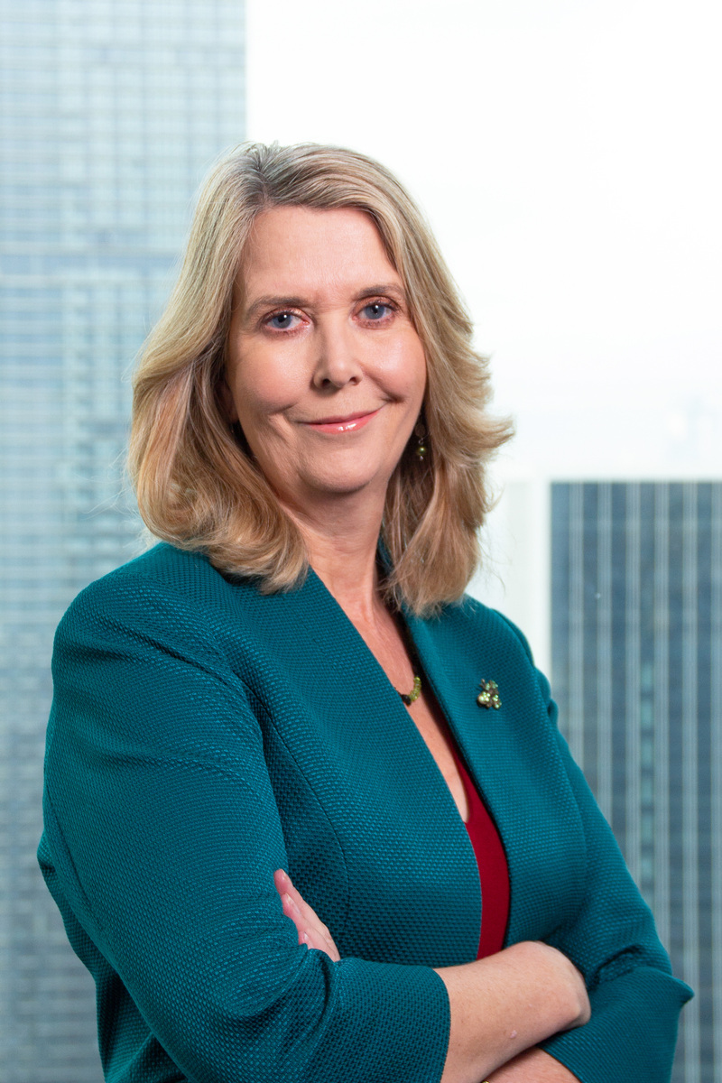 Smiling professional woman with blonde hair wearing a teal blazer over a red top, with office buildings in the background