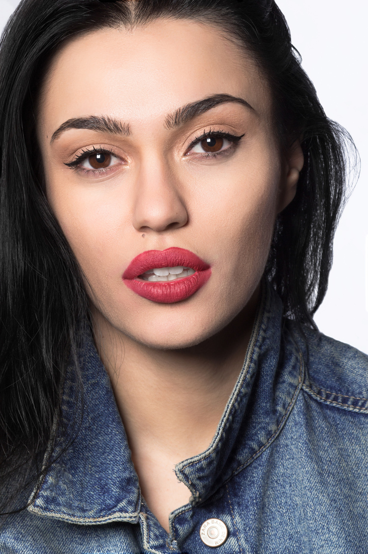 Close-up headshot of a young actress with striking red lipstick and black hair, wearing a denim jacket.